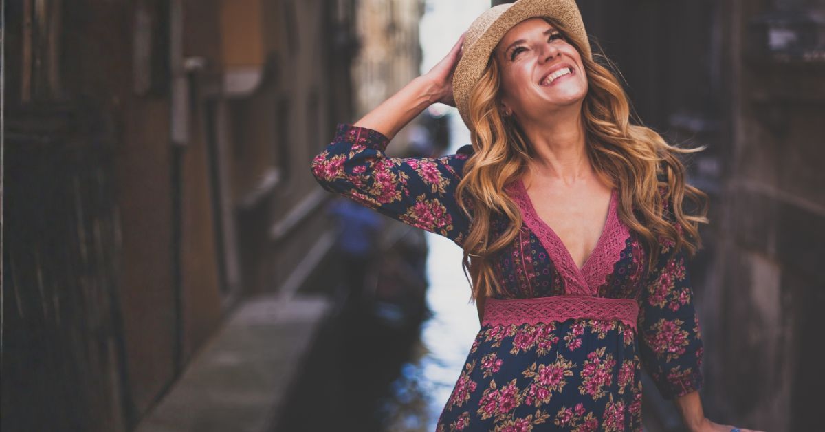 Consistency Woman in a floral dress and straw hat smiles as she stands in a narrow alleyway outdoors, body tilted back slightly as she enjoys the moment.