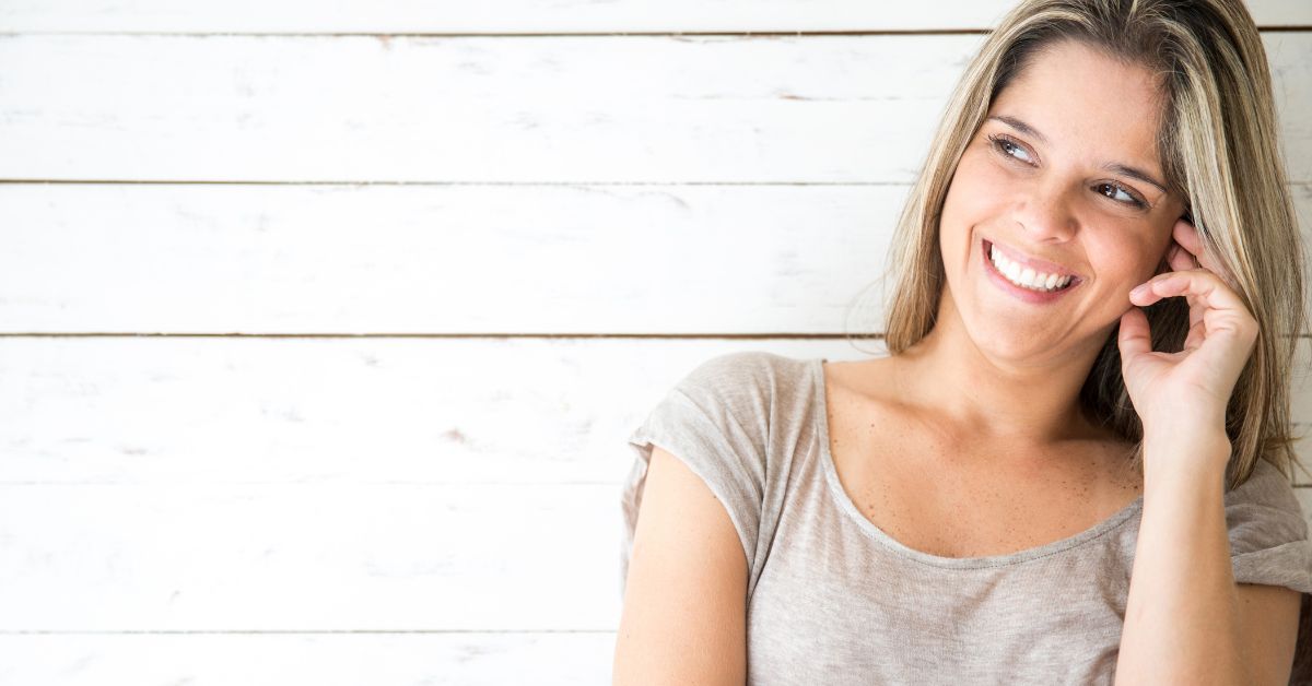 Smiling woman with blonde hair wearing a beige top, leaning against a white wooden plank background