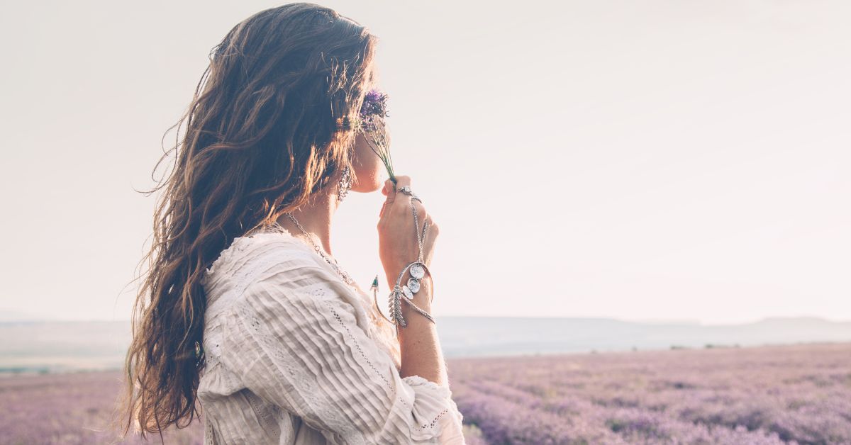 Woman with long wavy hair in a white blouse smelling a small purple flower in a lavender field at sunset/blooming field