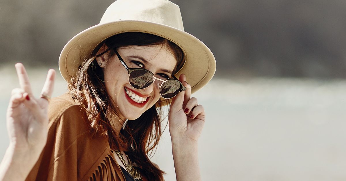 Smiling woman at the beach wearing a wide-brim hat and sunglasses, flashing a peace sign at the camera.